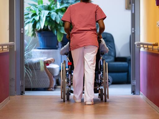 Eine Pflegeheimbewohnerin wird von einer Pflegerin mit dem Rollstuhl einen Gang entlanggeschoben.