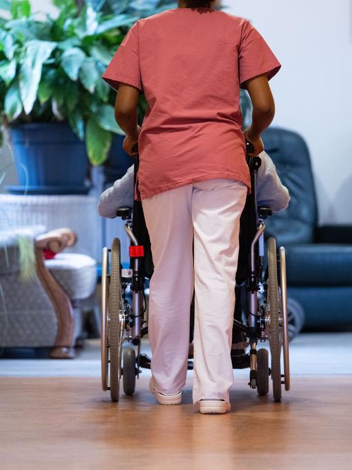 Eine Pflegeheimbewohnerin wird von einer Pflegerin mit dem Rollstuhl einen Gang entlanggeschoben.