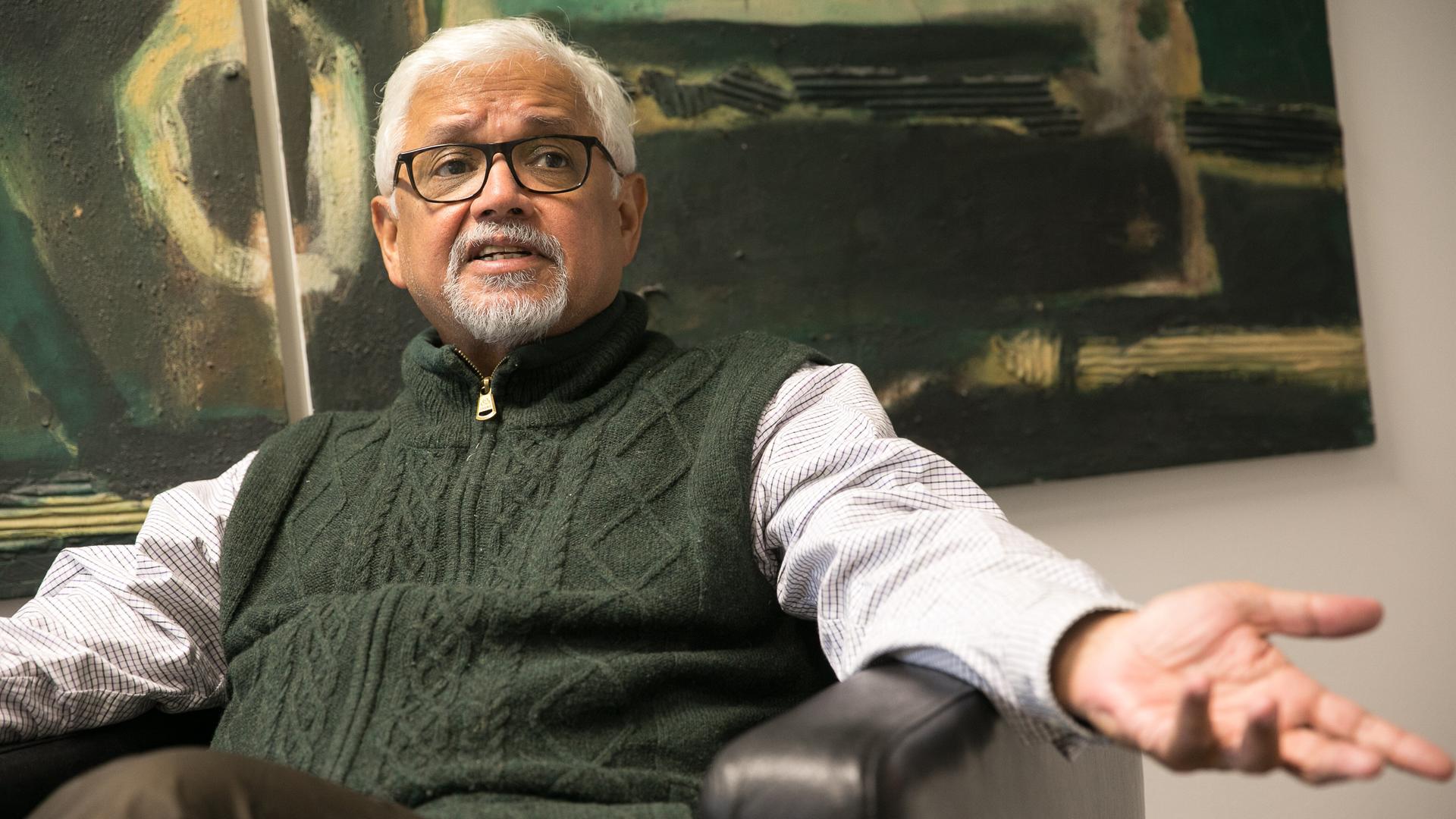 Amitav Ghosh sitzt im Sessel vor einem abstrakten Gemälde und ist angeregt im Gespräch.