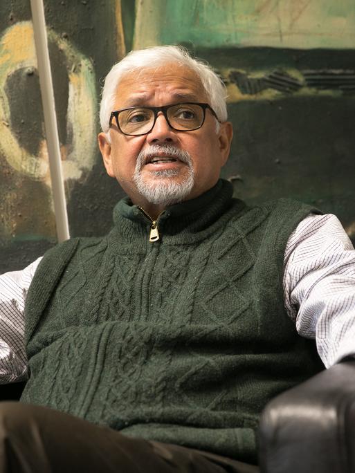 Amitav Ghosh sitzt im Sessel vor einem abstrakten Gemälde und ist angeregt im Gespräch.
