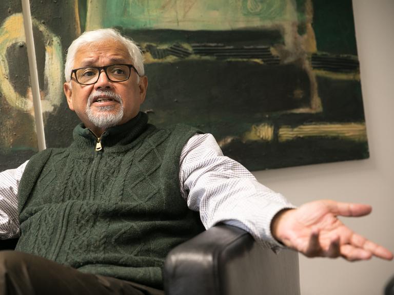 Amitav Ghosh sitzt im Sessel vor einem abstrakten Gemälde und ist angeregt im Gespräch.