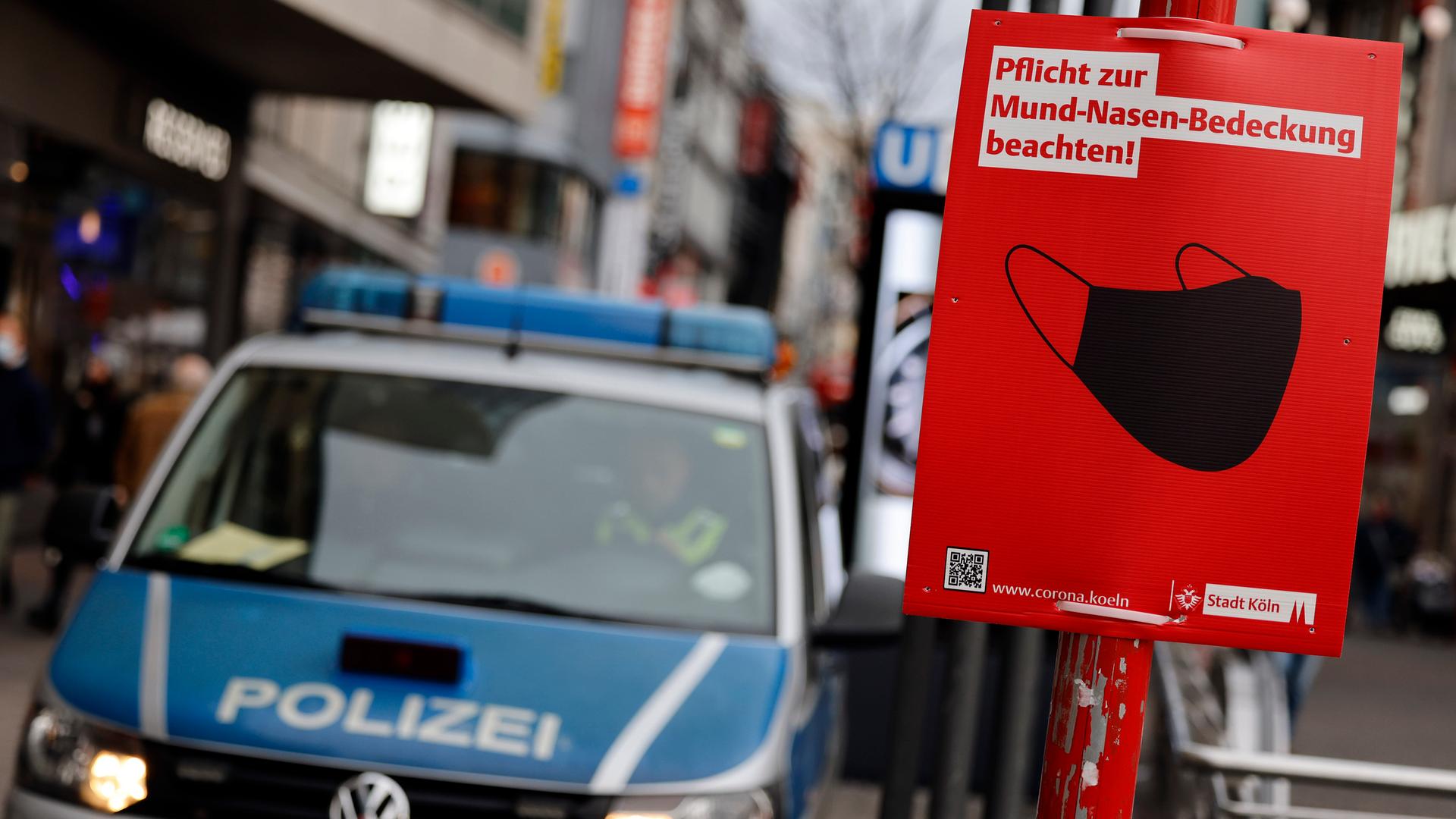 Ein Polizeiauto und ein Schild der Stadt Köln, auf dem auf die Maskenpflicht in der Öffentlichkeit während der Coronapandemie hingewiesen wird.