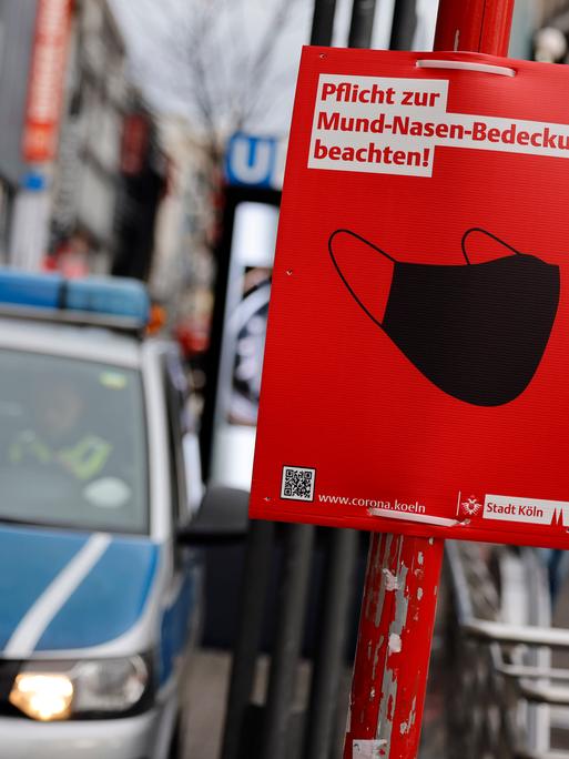 Ein Polizeiauto und ein Schild der Stadt Köln, auf dem auf die Maskenpflicht in der Öffentlichkeit während der Coronapandemie hingewiesen wird.