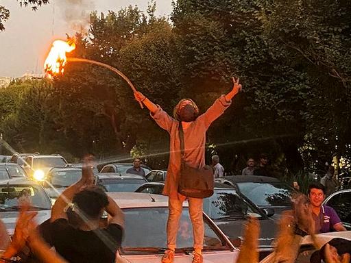 Eine Frau steht auf einem Auto bei Protesten in Teheran. Sie hat ihr Kopftuch abgelegt und die Arme zur Seite ausgestreckt.
