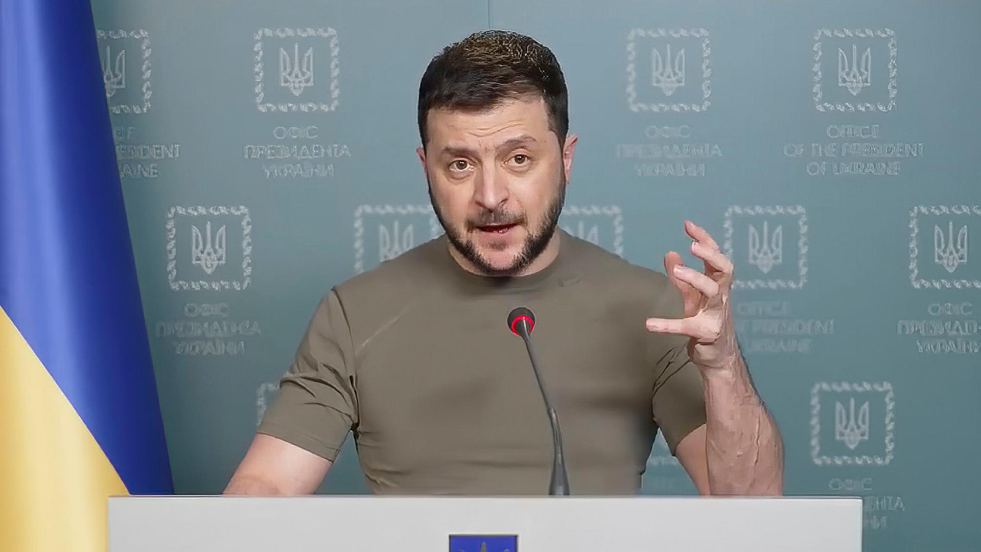 Der ukrainischen Präsident Selenskyj steht hinter einem Rednerpult, schaut in die Kamera und spricht in ein Mikrofon. Mit einer Hand gestikuliert er.