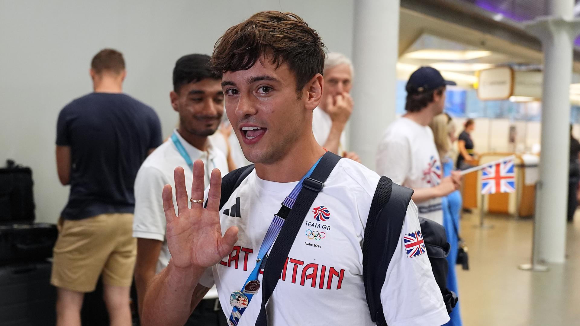 Tom Daley bei seiner Ankunft in London. Ein dunkelhaariger lächelnder Mann. Er trägt ein Shirt mit den Enblemen der Olympischen Spiele und Großbritanniens. Seine recht Hand ist erhoben. Tom Daley bei seiner Ankunft in London. Ein dunkelhaariger lächelnder Mann. Er trägt ein Shirt mit den Enblemen der Olympischen Spiele und Großbritanniens. Seine recht Hand ist erhoben.