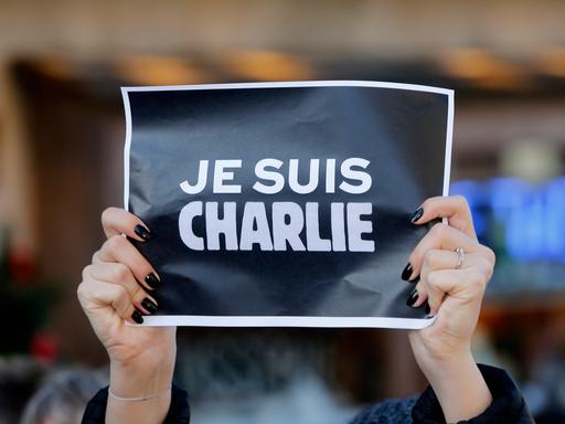 Weiße Schrift auf schwarzem Papier mit dem Aufdruck "Charlie Hebdo" gehalten bei einer Hand mit schwarzem Nagellack. Weiße Schrift auf schwarzem Papier mit dem Aufdruck "Charlie Hebdo" gehalten bei einer Hand mit schwarzem Nagellack.