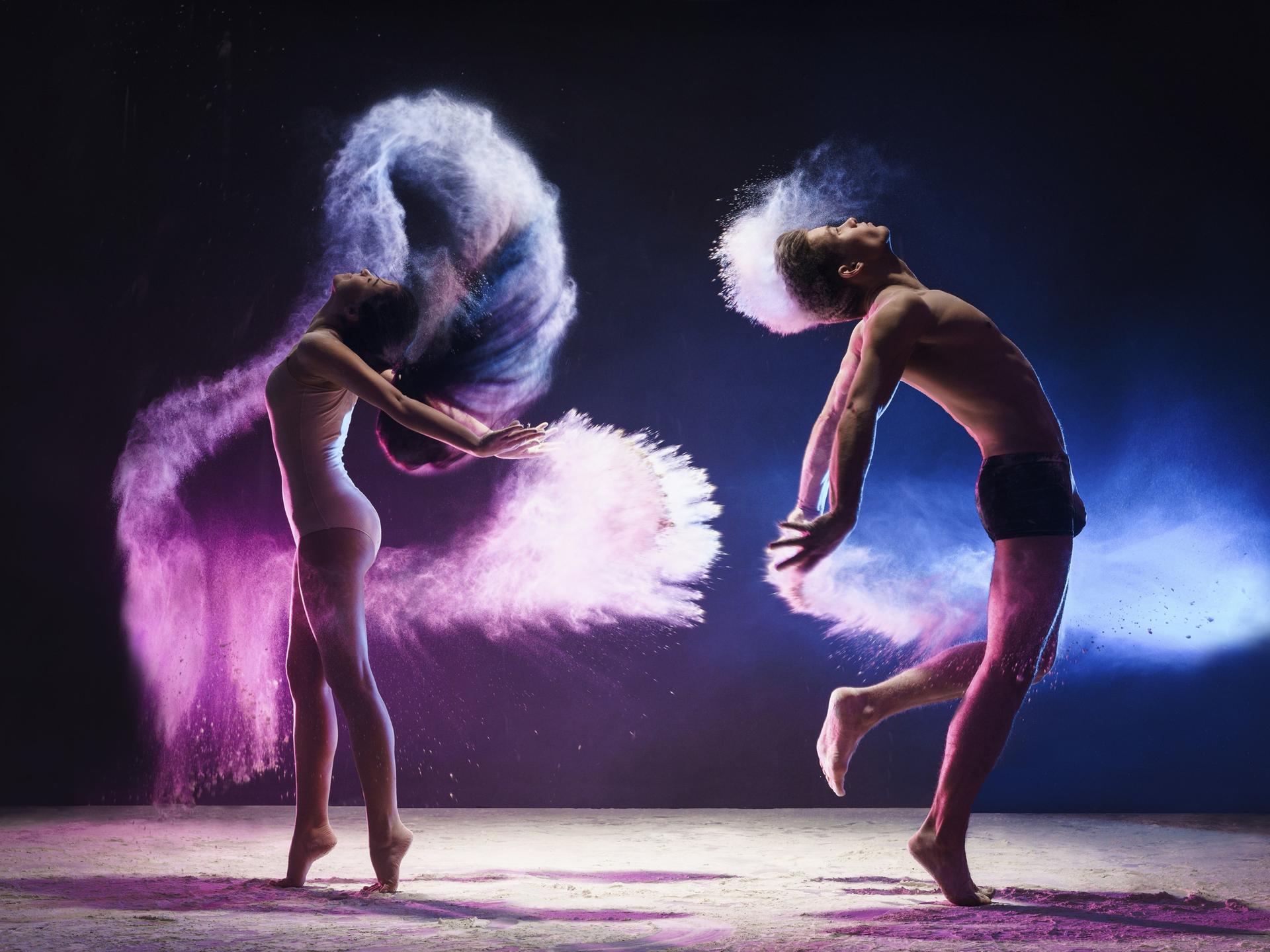 
Ein junger Mann und eine junge Frau sind mitten im tänzerischen Sprung aufgenommen, sie springen in einer pinken und blauen Wolke im Studio
