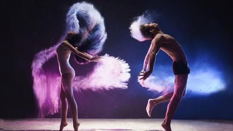 
Ein junger Mann und eine junge Frau sind mitten im tänzerischen Sprung aufgenommen, sie springen in einer pinken und blauen Wolke im Studio