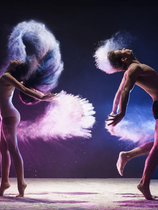 
Ein junger Mann und eine junge Frau sind mitten im tänzerischen Sprung aufgenommen, sie springen in einer pinken und blauen Wolke im Studio