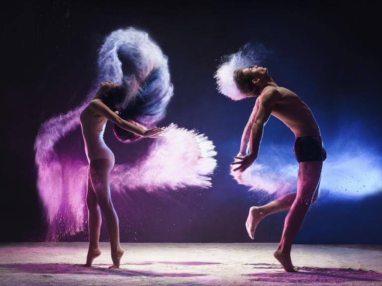 
Ein junger Mann und eine junge Frau sind mitten im tänzerischen Sprung aufgenommen, sie springen in einer pinken und blauen Wolke im Studio