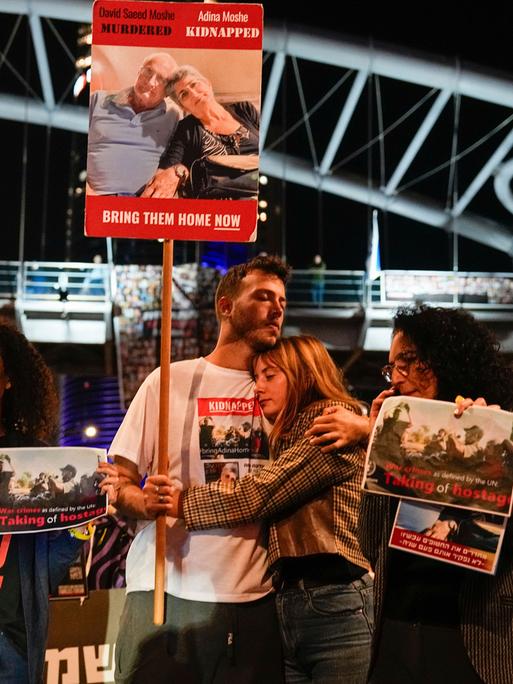 Menschen halten Plakate ihrer Angehörigen hoch. Ein Paar steht in der Mitte des Bildes und umarmt sich.