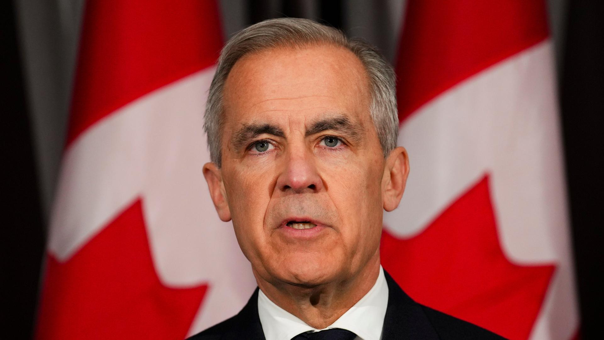 Der kanadische Premierminister Mark Carney steht im dunklen Anzug vor kanadischen Fahnen. 