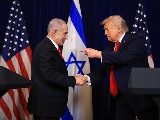 Benjamin Netanjahu und Donald Trump am 29.12.2025 in Trumps Privatresidenz Mar-a-lago in Florida. Sie schütteln Hände und Trump zeigt mit dem Finger der anderen Hand auf Netanjahu.