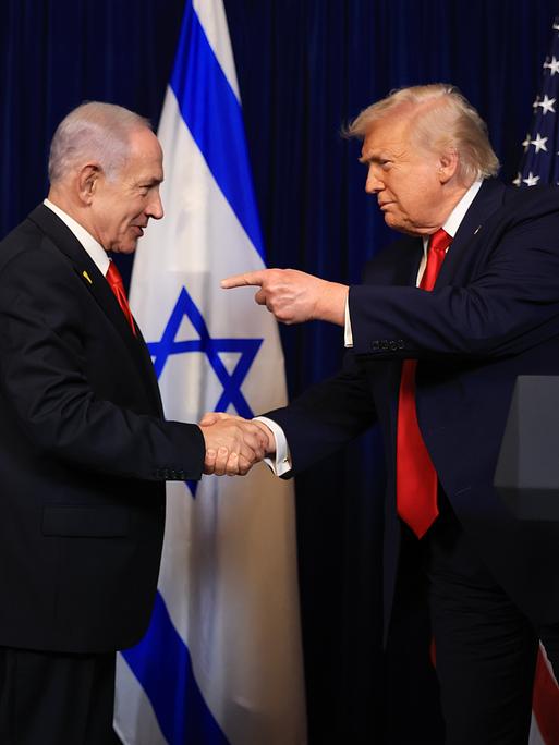 Benjamin Netanjahu und Donald Trump am 29.12.2025 in Trumps Privatresidenz Mar-a-lago in Florida. Sie schütteln Hände und Trump zeigt mit dem Finger der anderen Hand auf Netanjahu.