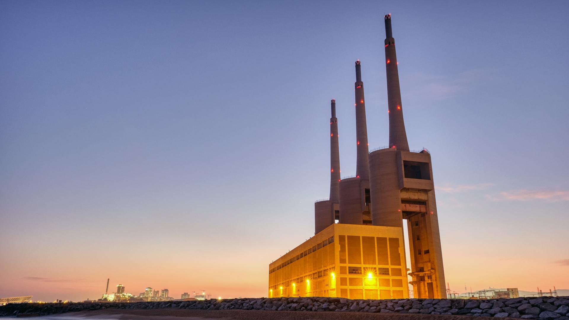 Zu sehen ist ein ehemaliges Heizkraftwerk in Barcelona am Strand. Das Kraftwerk wird dominiert von drei riesigen Schornsteinen. 