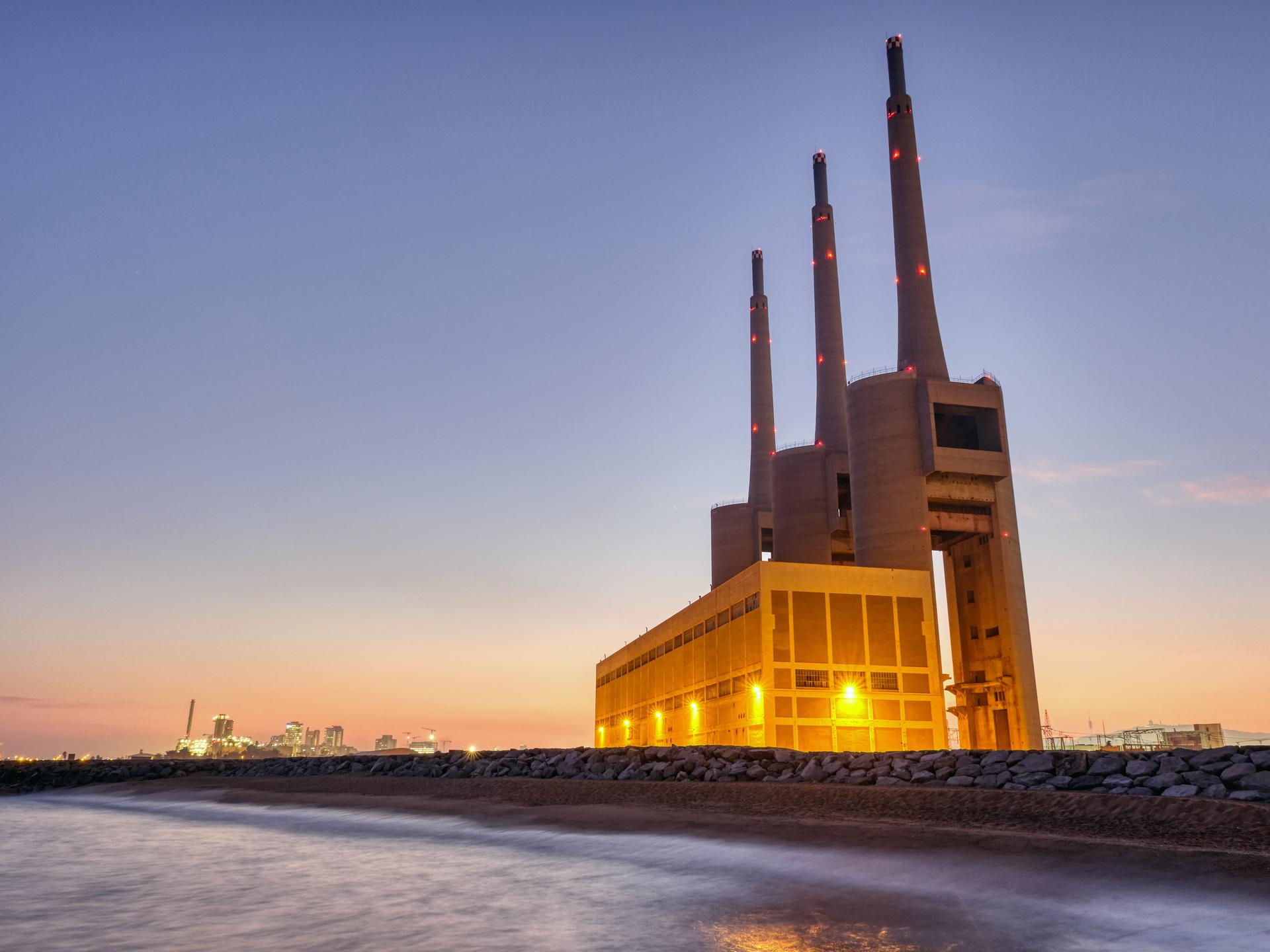 Zu sehen ist ein ehemaliges Heizkraftwerk in Barcelona am Strand. Das Kraftwerk wird dominiert von drei riesigen Schornsteinen. 