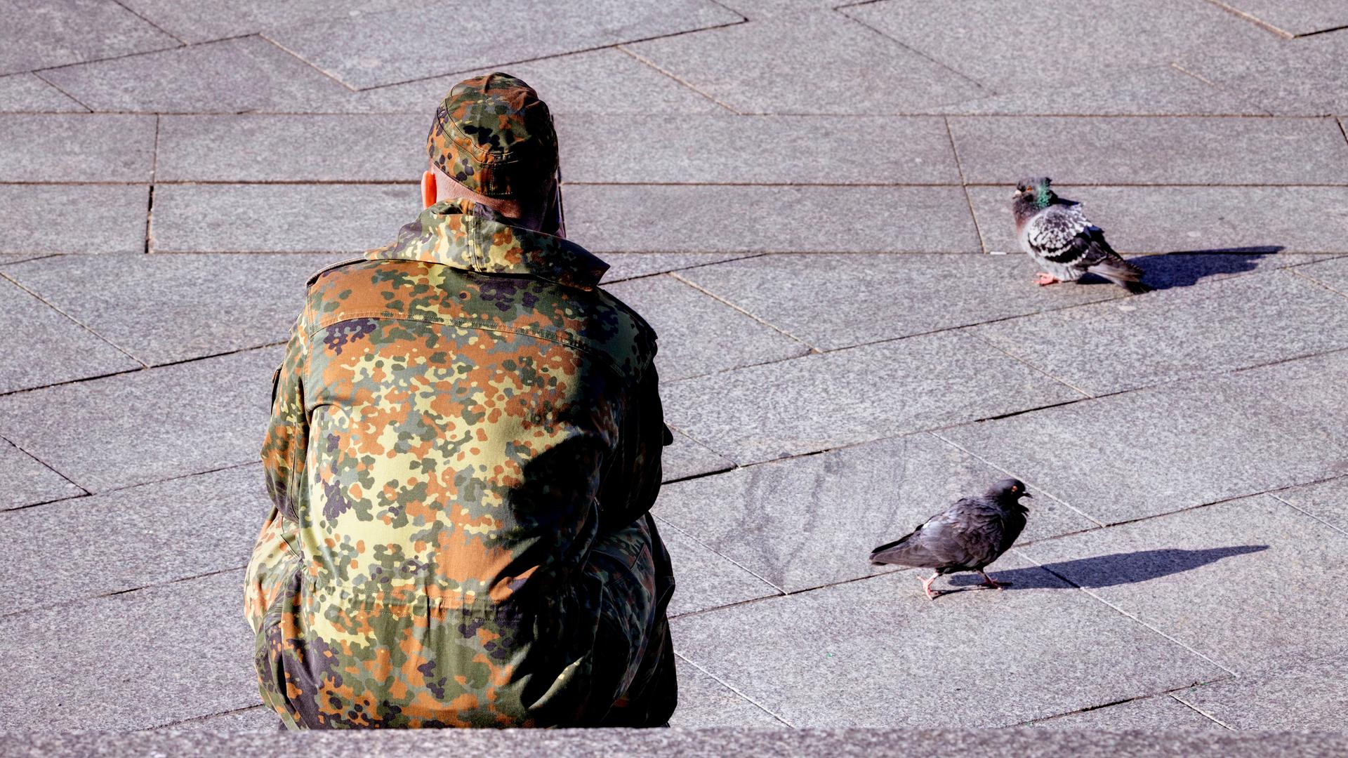 Ein von hinten fotografierter Bundeswehrsoldat sitzt auf einer Steintreppe, neben ihm zwei Tauben. 
