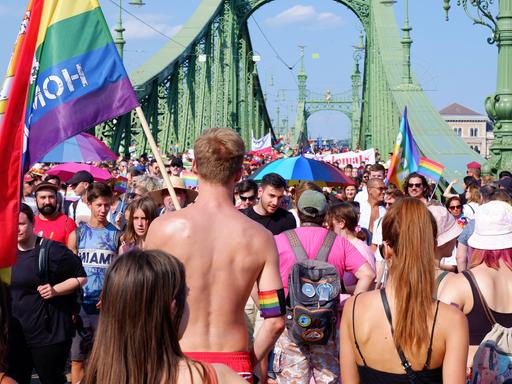Teilnehmende der Pride-Parade gehen in Budapest über ein Brücke. Ein Mann geht mit nacktem Oberkörper und trägt eine bunte Pride-Flaggen.