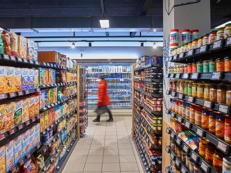 Eine Kundin im roten Mantel läuft zwischen Regalen in einem Supermarkt umher.