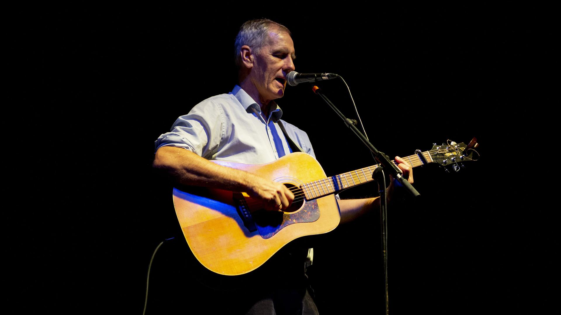 Der australische Singer-Songwriter, Gittarist und Musikkritiker, Robert Forster, steht im November 2019 in Porto mit der Gitarre umgehängt vor einem Mikrofon auf einer Bühne, der Hintergrund ist schwarz.