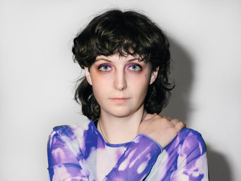 Porträt von Fiona Sironic. Sie trägt ein lila gemustertes Oberteil und hat schulterlanges, dunkelbraunes Haar.