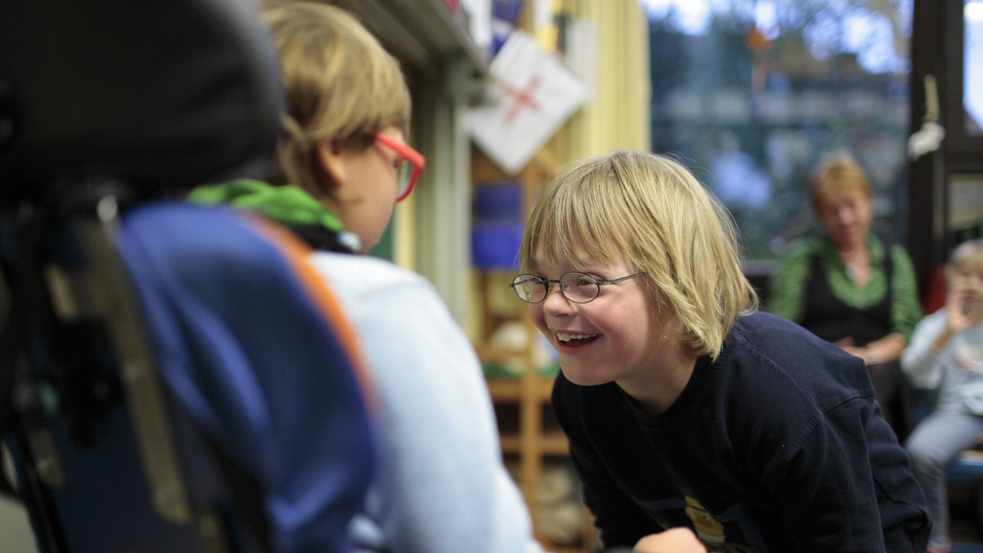 Ein Kind mit Down-Syndrom beugt sich lachend zu einem Kind im Roll-Stuhl vor. 