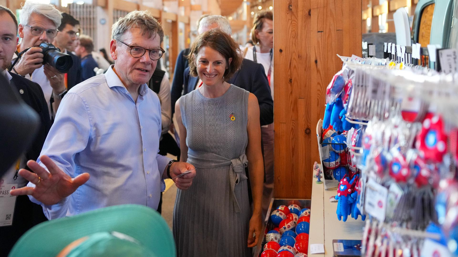 Wadephul im blauen Hemd steht vor einem Souvenirstand , sagt etwas und gestikuliert mit der linken Hand. Daneben eine lächelnde Frau und weitere Personen, darunter ein Fotograf. Wadephul im blauen Hemd steht vor einem Souvenirstand , sagt etwas und gestikuliert mit der linken Hand. Daneben eine lächelnde Frau und weitere Personen, darunter ein Fotograf.