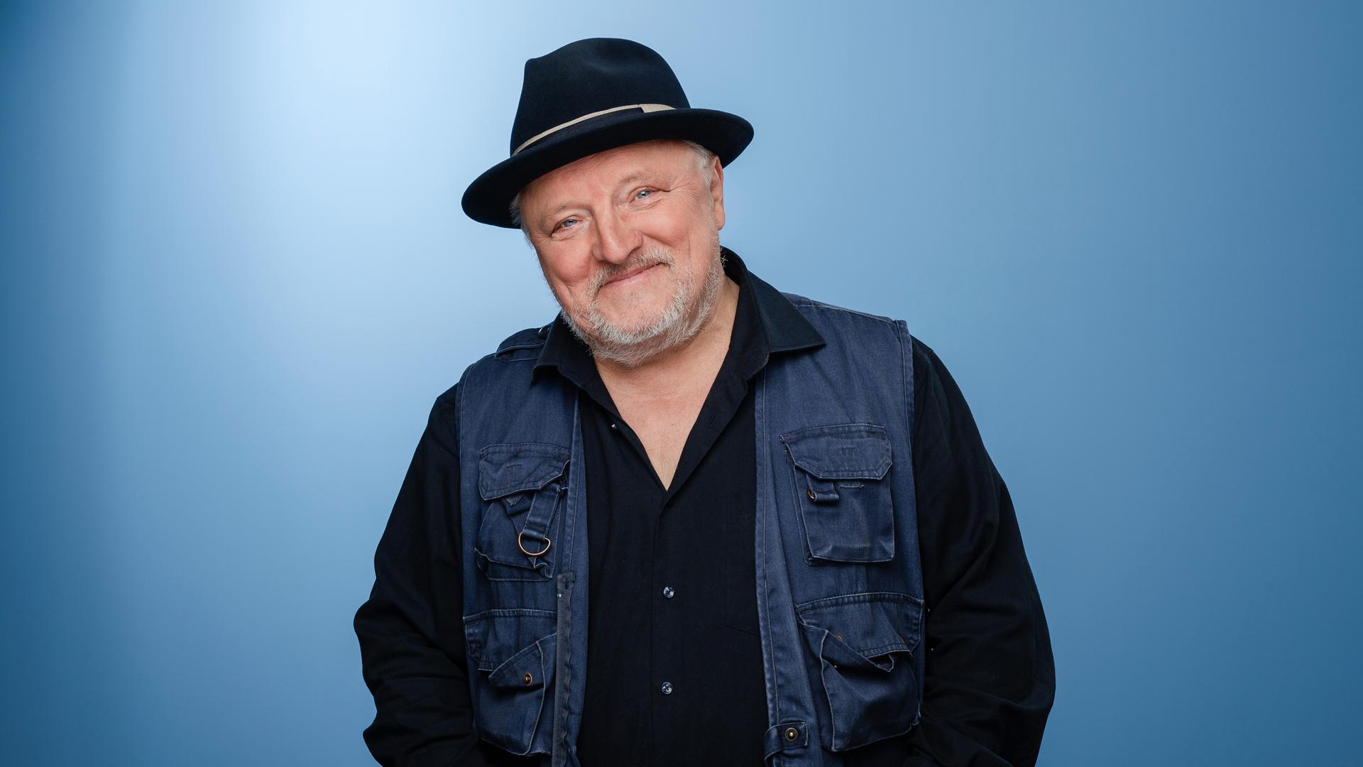 Axel Prahl trägt Dreitagebart, dunkle Kleidung und einen schwarzen Hut. Er steht in einem Fotostudio mit blauem Hintergrund und lacht in die Kamera. Axel Prahl trägt Dreitagebart, dunkle Kleidung und einen schwarzen Hut. Er steht in einem Fotostudio mit blauem Hintergrund und lacht in die Kamera.