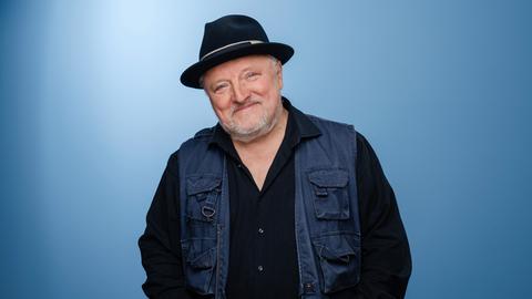 Axel Prahl trägt Dreitagebart, dunkle Kleidung und einen schwarzen Hut. Er steht in einem Fotostudio mit blauem Hintergrund und lacht in die Kamera. Axel Prahl trägt Dreitagebart, dunkle Kleidung und einen schwarzen Hut. Er steht in einem Fotostudio mit blauem Hintergrund und lacht in die Kamera.
