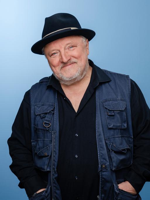 Axel Prahl trägt Dreitagebart, dunkle Kleidung und einen schwarzen Hut. Er steht in einem Fotostudio mit blauem Hintergrund und lacht in die Kamera.