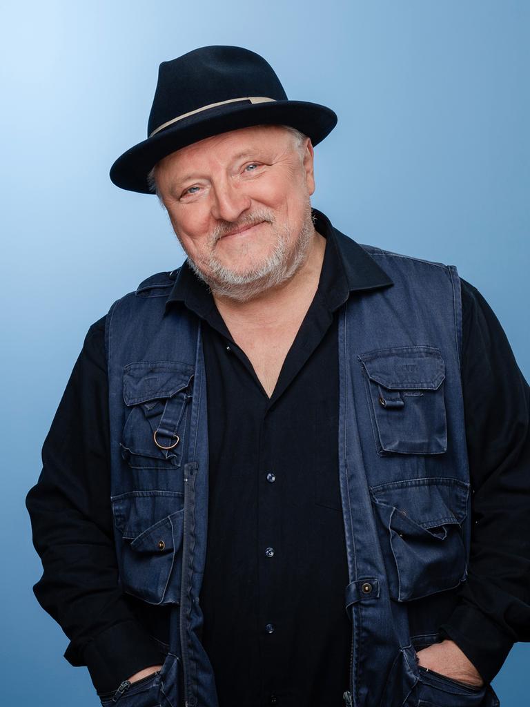 Axel Prahl trägt Dreitagebart, dunkle Kleidung und einen schwarzen Hut. Er steht in einem Fotostudio mit blauem Hintergrund und lacht in die Kamera.
