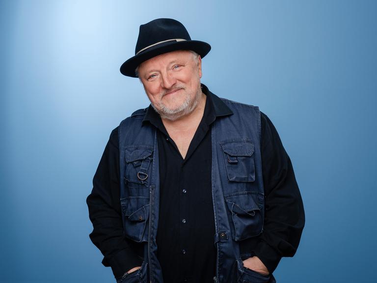 Axel Prahl trägt Dreitagebart, dunkle Kleidung und einen schwarzen Hut. Er steht in einem Fotostudio mit blauem Hintergrund und lacht in die Kamera.