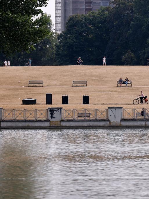 Blick auf einen Park mit komplett vertrockneter Wiese. Im Vordergrund ein Wasserbecken, im Hintergrund vereinzelt Menschen auf Bänken, beim Radfahren oder joggen.