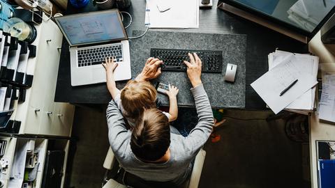 Eine Mutter sitzt mit einem Kind auf dem Schoß am Schreibtisch und versucht an einem Computer zu arbeiten.