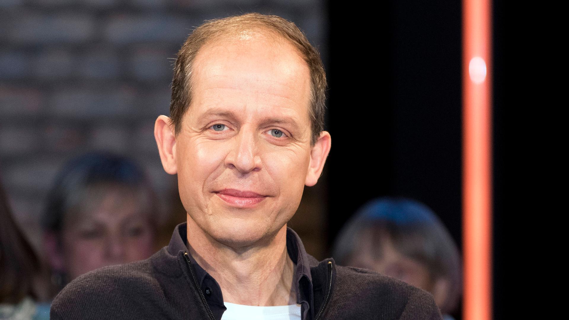 Stephan Schäfer sitzt im Studio bei einer TV-Aufzeichnung. Er trägt ein weißes T-Shirt und darüber einen schwarzen Pullower. Er guckt in die Kamera. Im Hintergrund ist verschwommen Publikum zu erkennen.