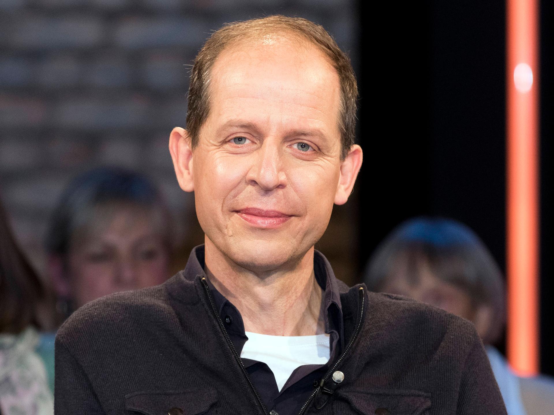 Stephan Schäfer sitzt im Studio bei einer TV-Aufzeichnung. Er trägt ein weißes T-Shirt und darüber einen schwarzen Pullower. Er guckt in die Kamera. Im Hintergrund ist verschwommen Publikum zu erkennen.