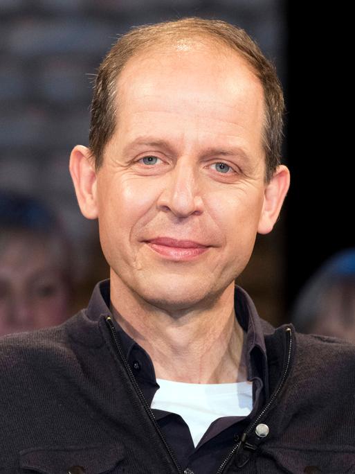 Stephan Schäfer sitzt im Studio bei einer TV-Aufzeichnung. Er trägt ein weißes T-Shirt und darüber einen schwarzen Pullower. Er guckt in die Kamera. Im Hintergrund ist verschwommen Publikum zu erkennen.