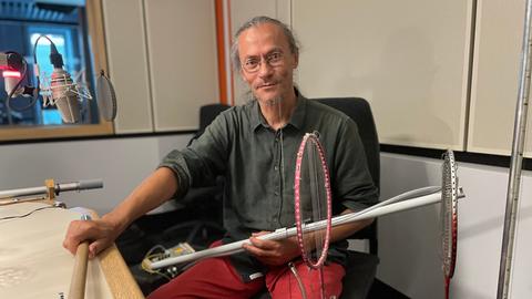 Porträt von Ardhi Engl im Deutschlandradio Studio. Er hat sein graues Haar zum Zopf gebunden, trägt eine Brille und hält mehrere obskure Instrumente in den Händen.