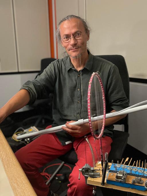 Porträt von Ardhi Engl im Deutschlandradio Studio. Er hat sein graues Haar zum Zopf gebunden, trägt eine Brille und hält mehrere obskure Instrumente in den Händen.
