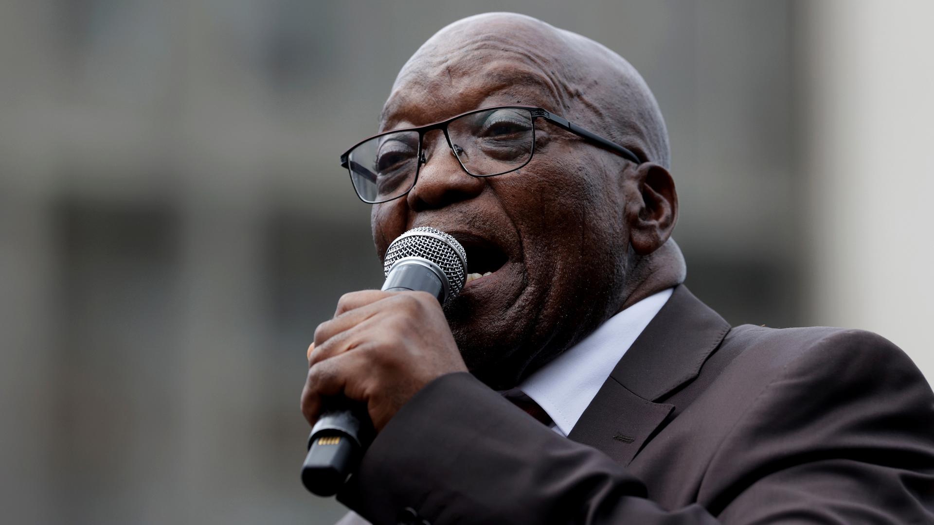 Zuma im dunklen Jackett und mit Brille spricht engagiert in ein Mikrofon.