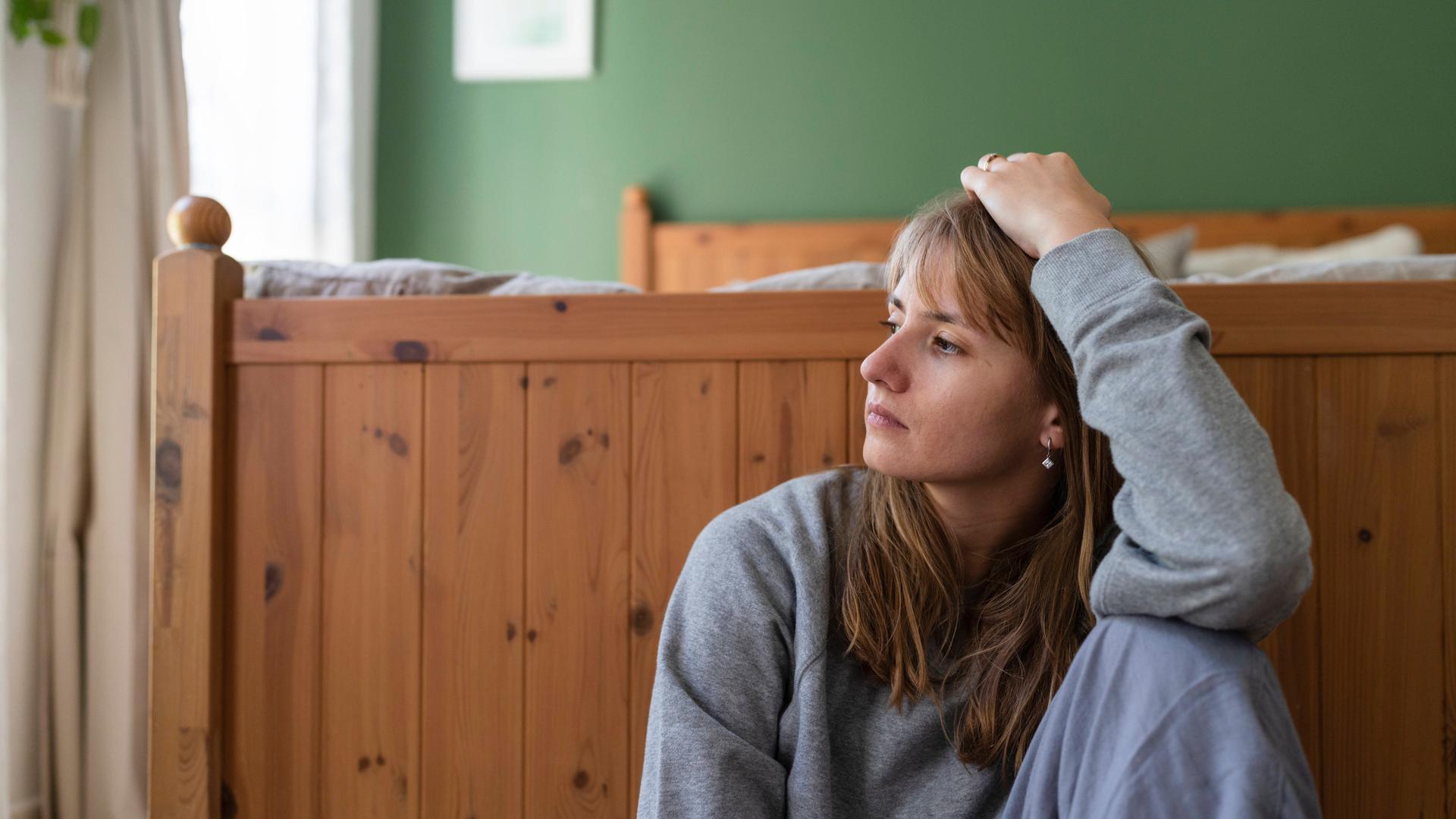Eine junge Frau sitzt nachdenklich auf dem Boden vor einem Bett