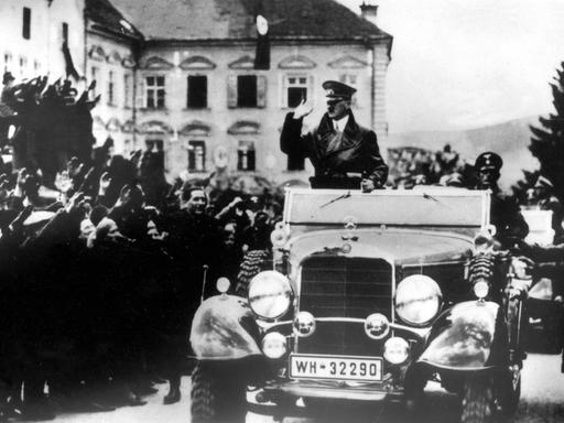 Historische Schwarz-weiß-Aufnahme zeigt Adolf Hitler im offenen Fahrzeug. Ringsherum stehen zahlreiche Menschen und jubeln ihm zu.