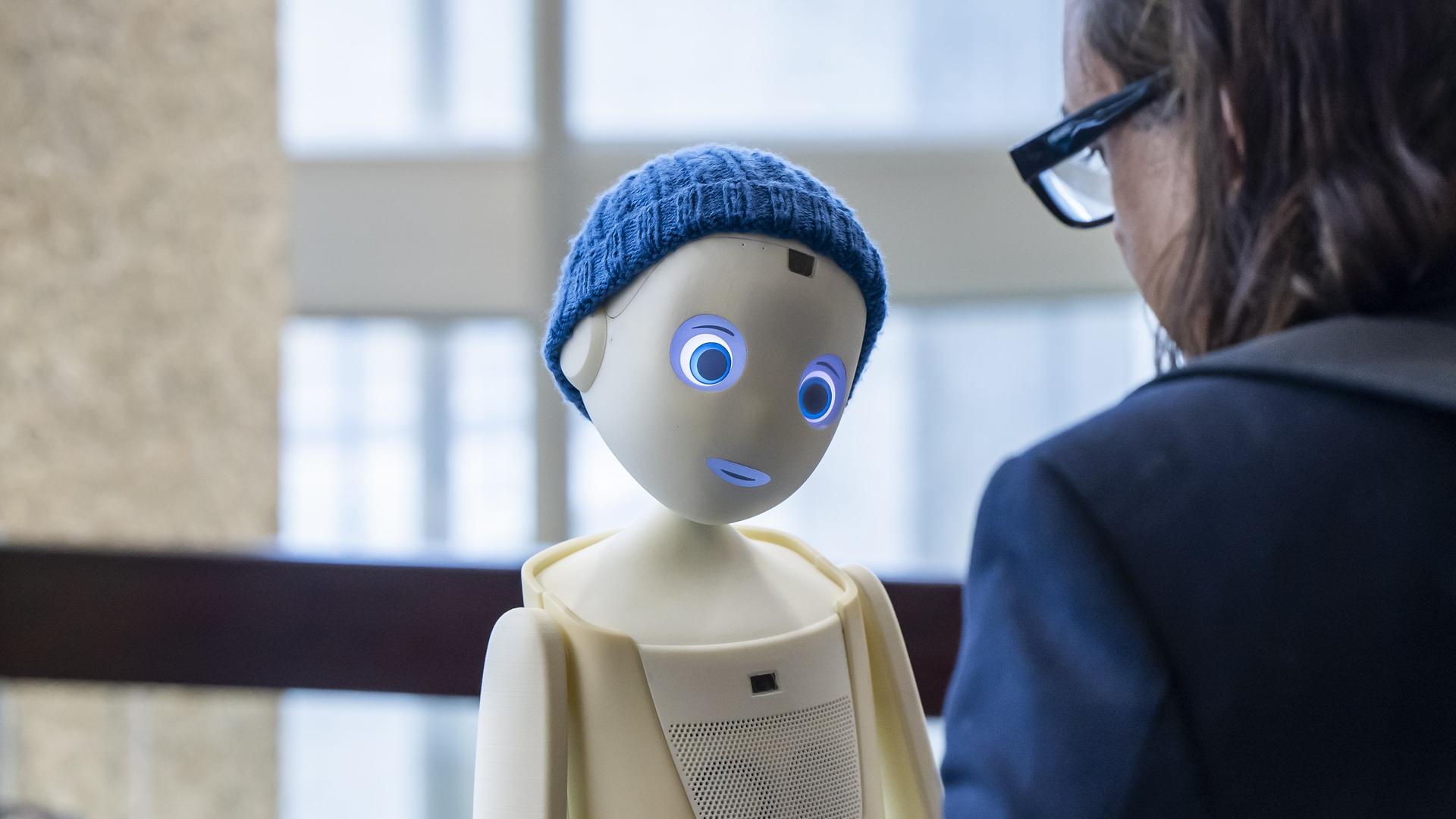Der Sozialroboter Oscar der Münchner Firma Navel Robotics im Bildzentrum, mit Strickmütze und großen Augen, ihm gegenüber eine Frau in Rückansicht