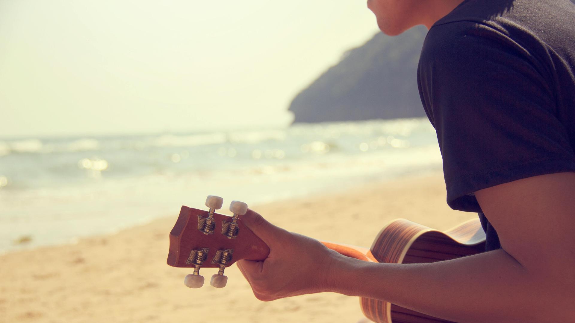 Blick von hinten auf einen Menschen, der am Meer Ukulele spielt.
