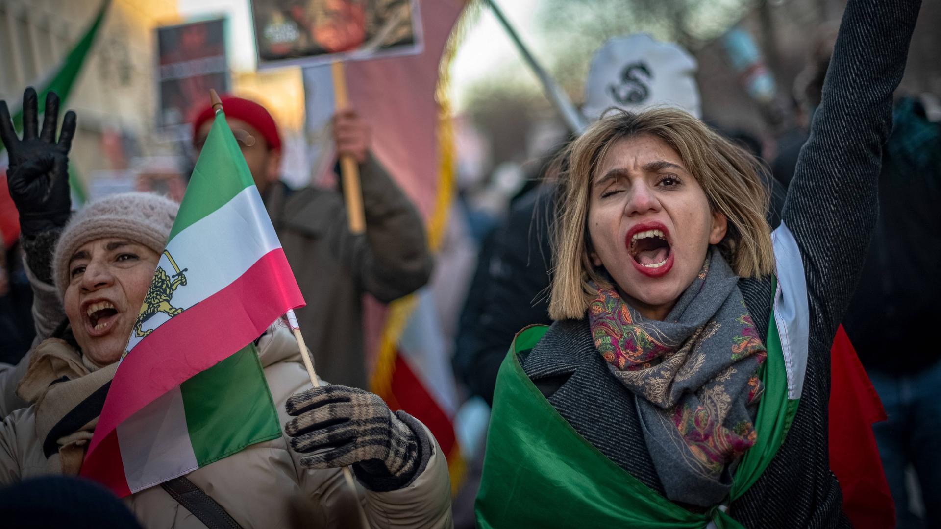 Zwei Frauen demonstrieren zusammen mit anderen Menschen. Sie tragen iranische Flaggen, rufen und heben dabei die Hände.
