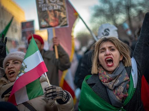 Zwei Frauen demonstrieren zusammen mit anderen Menschen. Sie tragen iranische Flaggen, rufen und heben dabei die Hände. Zwei Frauen demonstrieren zusammen mit anderen Menschen. Sie tragen iranische Flaggen, rufen und heben dabei die Hände.