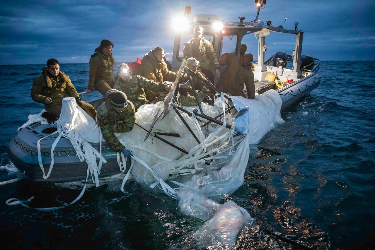 Mehrere Soldaten in Tarnuniformen sitzen in einem kleinen Boot. Einige von ihnen ziehen Überreste der weißen Ballonhülle ins Boot, dessen beide Scheinwerfer in die Kamera strahlen. Es ist Abend- oder Morgendämmerung.  