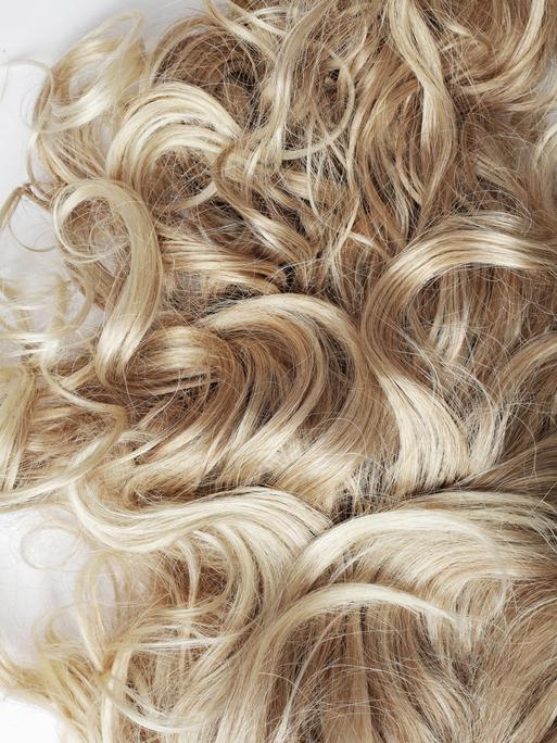 Ein Detailfoto von blonden, gelocktem Haar. Es scheint auf einem Tisch oder einem anderen weißen Untergrund zu liegen.