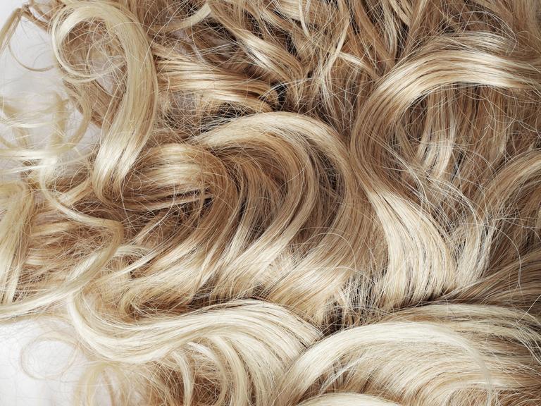Ein Detailfoto von blonden, gelocktem Haar. Es scheint auf einem Tisch oder einem anderen weißen Untergrund zu liegen.